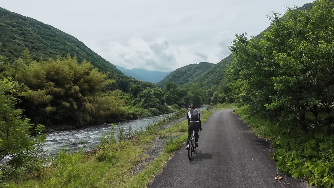 A rider on a quiet paved road beside the rushing Kiso River, green hills on both sides